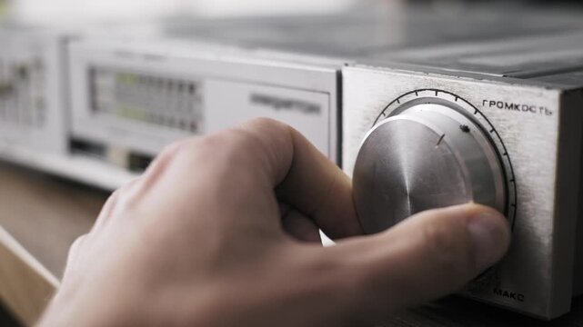 Close-up of a male hand adjusting the round volume knob on a vintage Soviet-era audio amplifier. The scratched silver metal faceplate evokes a sense of nostalgia for analog sound.