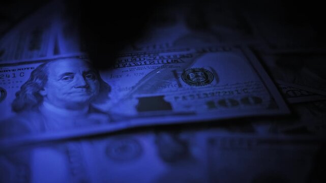 Macro view of an expert inspecting a one hundred dollar bill using a portable UV light detector in darkness. The blue light verifies hidden security features to check for forgery.