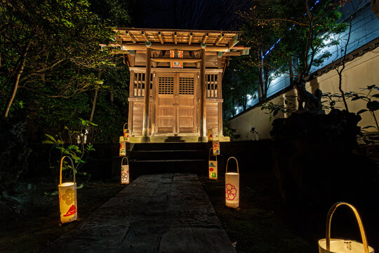 Shinto shrine building and stone fox statue with glowing lanterns at Koishikawa Korakuen at night January 2024