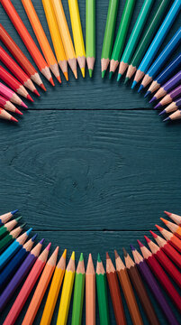 A vibrant spectrum of colored pencils forms a circular border on a textured dark blue wooden surface, inviting creativity.