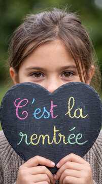 A young girl with brown hair holds a heart-shaped slate with "C'est la rentr&eacute;e" written in colorful chalk.