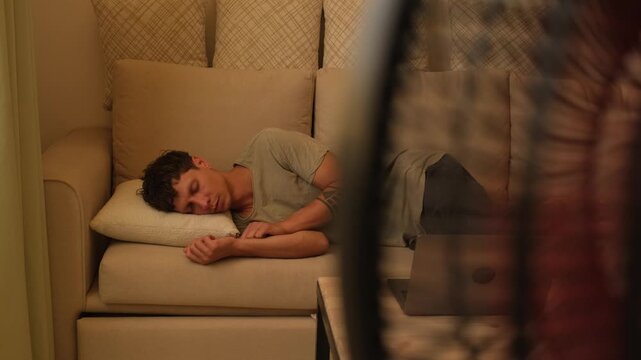 Living without air conditioning during extreme heat, exhausted young man resting on sofa near electric fan while trying to cope with high indoor temperature