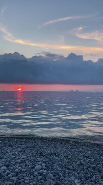 orange sun sinks beneath a dramatic dark cloud bank above a calm sea, casting a long glowing reflection across gentle waves beside a quiet pebble beach at twilight