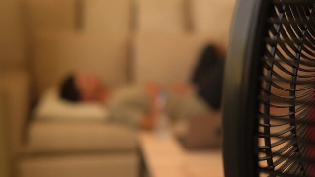 Living without air conditioning during extreme heat, electric fan in focus with exhausted man resting in blurred background, copy space for text and climate related message
