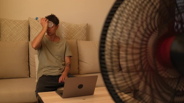 Young man struggling with extreme heat at home, working on laptop, cooling forehead with cold water bottle and drinking water during heatwave indoors