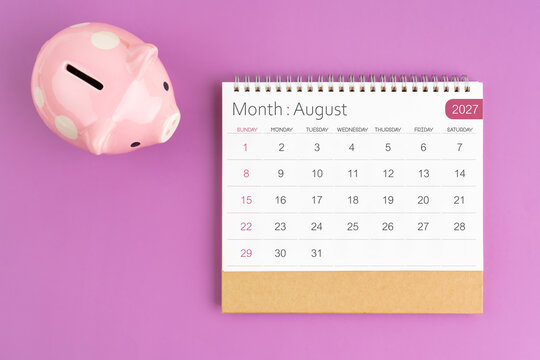 August 2027 desk calendar and pink piggy bank on purple background.