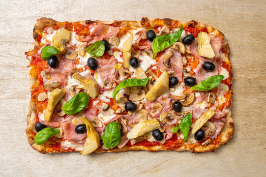 Traditional Roman-style quattro stagioni pinsa topped with tomato sauce, mozzarella, cooked ham, mushrooms, artichokes, black olives and fresh basil, photographed from above on a rustic wooden backgro
