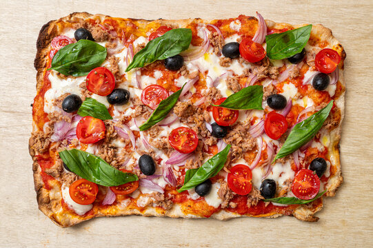 Traditional Roman-style pinsa topped with tomato sauce, mozzarella, tuna, red onion, black olives, cherry tomatoes and fresh basil, photographed from above on a rustic wooden background.