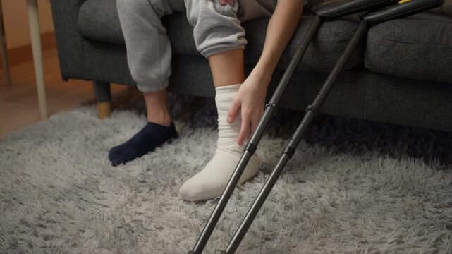 Close-up of  teenager boy  with bandaged leg sitting on sofa at home, gently massaging injured ankle. Crutches lean against couch, symbolizing recovery process from an accident
