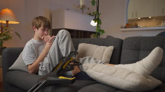 Smiling teenage boy with cast on broken leg rests on couch at home, happily browsing smartphone while crutches lean against sofa, symbolizing recovery and modern leisure