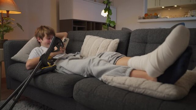 Teenage boy recovering from foot injury rests at home with bandaged leg elevated, smiling while using smartphone as crutches stand nearby during rehabilitation process.