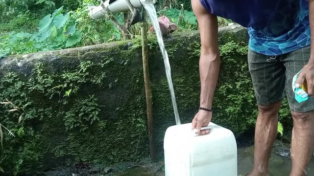 Man filling plastic jerrycan container with clean water near blue drum barrel outdoor
