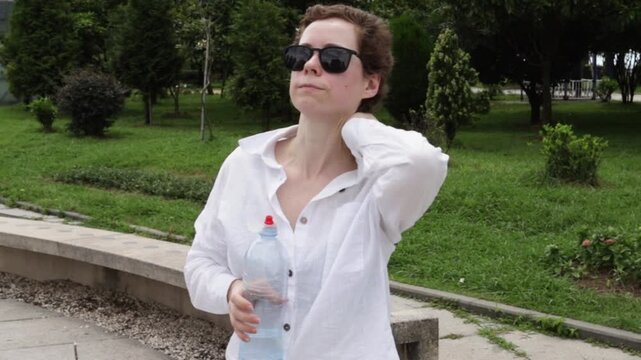 woman recovering from cancer holds a water bottle and cools her neck in a city park, illustrating overheating prevention, hydration routine and summer health awareness