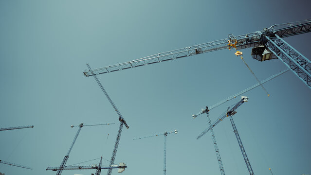 Dramatic low-angle view of multiple tower cranes crossing a clear blue sky, illustrating heavy construction, engineering scale and fast-paced urban expansion