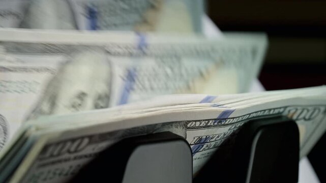 Automatic money counting machine processes stacks of US dollar bills illustrating inflation, purchasing power, rising prices, economic uncertainty and financial markets.
