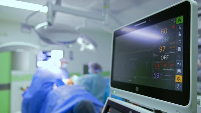 Distancing from the lung ventilation machine monitor. Medics work at the operating table in the blurred backdrop.