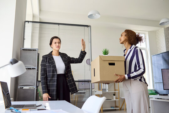 Employee and manager in office during dismissal. Fired businesswoman holds a box as a stern colleague signals stop, illustrating termination or resignation . Workplace conflict and exit concept.