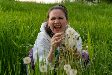 A laughing adult woman holds common dandelion stems, Taraxacum officinale, as numerous white seeds float around her in a green meadow. The expressive portrait captures joy, spontaneity, springtime fun © Volha