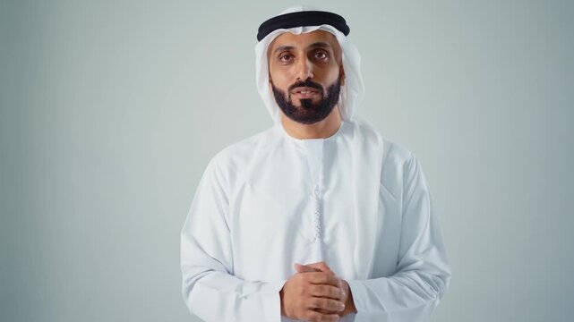 Confident young arab man with a beard wearing traditional clothing, a keffiyeh and agal, looking at the camera and smiling, expressing happiness and success on a gray studio background