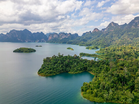 Aerial panoramic view of Cheow Lan Lake on quiet morning with evergreen lush trees and islands in the foreground and towering limestone mountains further back in Khao Sok National Park, Thailand