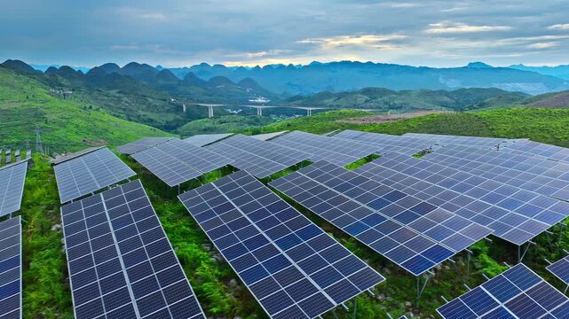 Solar panels on a green hill slope generating clean energy, with a scenic mountain range and bridge background under a cloudy sky.