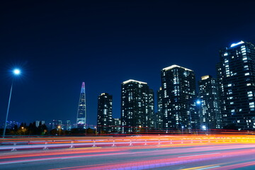 Fototapeta premium SEOUL, SOUTH KOREA - JUN 8, 2019: The distant Lotte World Tower stands tall beyond a row of modern residential skyscrapers and fast-moving traffic light trails along the highway at night.
