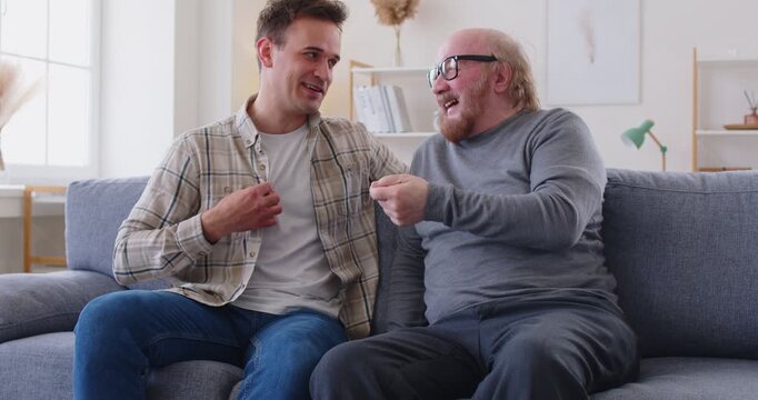 Senior father and adult son sitting on sofa in living room and having lively conversation. Cheerful smiling men enjoying their time, having fun and laughing during friendly family dialogue at home.