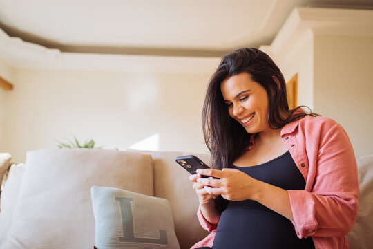 Smiling pregnant woman texting on smartphone at home sofa