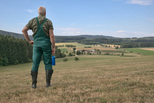 A farmer is standing on his sun-parched meadow in a hot summer, looking down into the valley where his farm is located. A symbol for agriculture and summer drought.