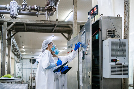 Worker in lab coat, hairnet, and gloves points at electrical panel. Holding tablet, they inspect controls in industrial setting. Pipes and machinery visible in factory environment