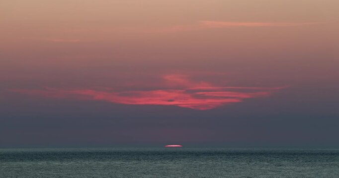 Tiny slither of the red pink setting sun disappears over the horizon sinking into the sea during midsummer 4K video shot in Cardigan Bay, Wales, United Kingdom, with a moody dramatic evening sky
