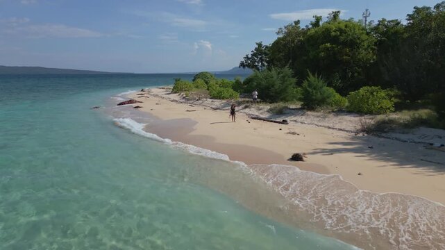 Low angle sweeping view of gentle waves splashing onto tropical beach with turquoise waters on Tabuhan Island with young woman walking in background, East Java