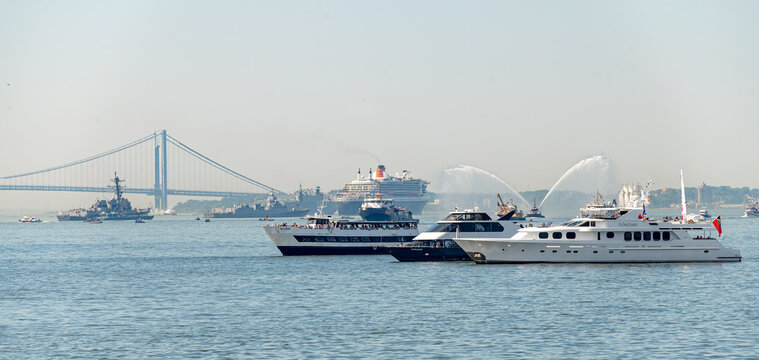 New York, USA, 4 Jule 2026 : Yachts gather before Queen Mary 2 during Independence Day. Passenger vessels line harbor water as fireboats spray arcs beside warships and Verrazzano Narrows Bridge.