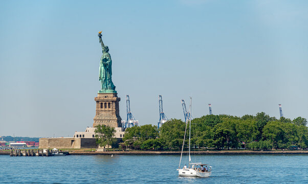 New York, USA, 4 Jule 2026 : Sailboat passes Statue of Liberty on New York Harbor. White yacht carries passengers over blue water before Liberty Island, green trees and distant port cranes.