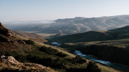 Obraz premium Mist drifts over the vast rugged mountain terrain where a river flows through a quiet green valley