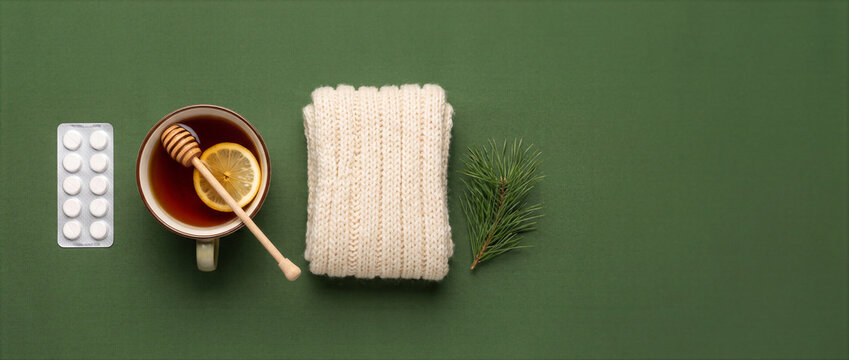Tea with lemon and honey, tablets, knitted scarf and pine branch on green background, winter cold prevention flat lay with copy space