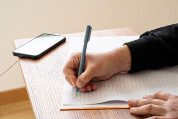 Fototapeta premium Man hand writing with blue pen on notebook page near smartphone