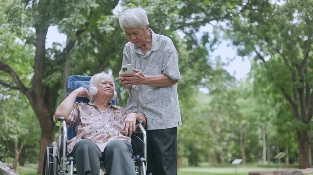 Smiling retired Asian mother with headphones chatting on a video call via smartphone, while her loving son stands by her side providing support in the beautiful summer park.