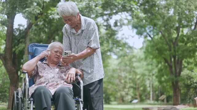 Joyful senior Asian mother in a wheelchair using a mobile phone and headphones to listen to music, laughing happily with her supportive adult son under lush green tree shade.