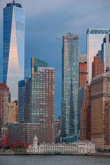 Fototapeta premium One World Trade Center above Lower Manhattan waterfront. Glass skyscrapers rise behind a white waterfront terminal under storm clouds at dusk.
