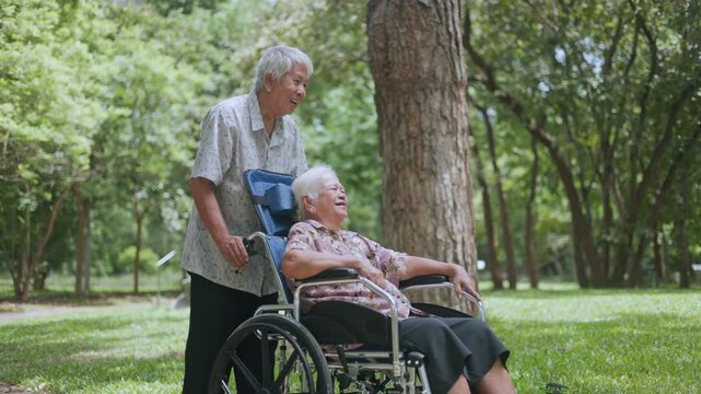 Smiling old Asian mother looking at her supportive son while being pushed in a wheelchair on a park trail, enjoying their high-quality time and healthy retirement lifestyle.