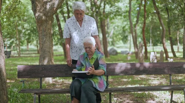 Loving elderly Asian couple enjoying a peaceful day in the park, husband standing and guiding his wife while she reads a memory book on a bench, sharing heartwarming moments outdoor.