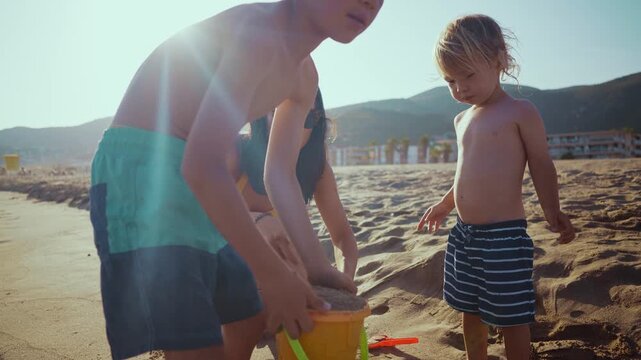 Beautiful family with a mother and two young sons playing with a bucket and shovel on a sandy beach during a sunny summer vacation, creating joyful childhood memories together by the sea