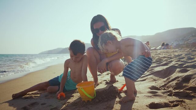 Beautiful family with a mother and two young sons playing with a bucket and shovel on a sandy beach during a sunny summer vacation, creating joyful childhood memories together by the sea