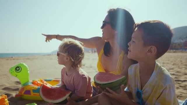 Cheerful mother with two sons sitting on a sandy beach, laughing together and eating a fresh watermelon during a sunny summer vacation, creating joyful and memorable family moments