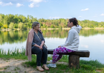 An elderly woman and younger adult woman sit face to face on a wooden bench beside a calm lake with common reed and green woodland. The image conveys intergenerational conversation, companionship, lis © Volha