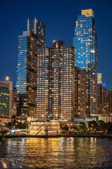 Fototapeta premium New York, USA, 8 June 2026 : Vertical view of New York City skyscrapers illuminated at night. Lit windows of modern apartment buildings reflect yellow light on the river surface, bottom up shot.