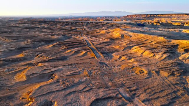 High angle aerial view of rugged desert yardang landform with vehicle tracks in Xinjiang, China. Captured at sunset with warm light reflecting on textured terrain.