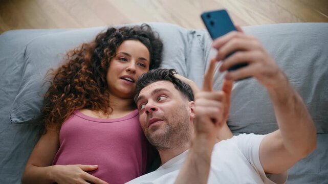 Happy couple taking selfie phone bed. Multiethnic pair browsing smartphone