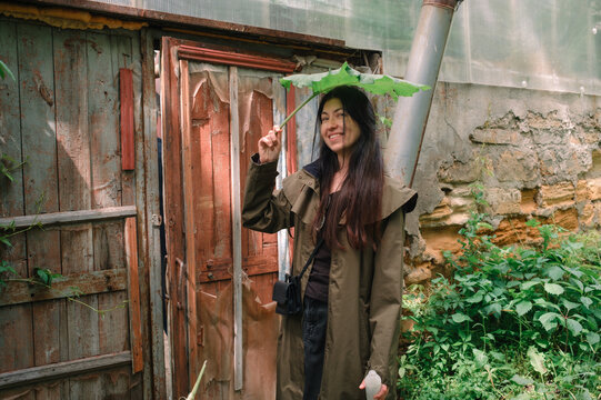 Woman holding large leaf as umbrella in garden near greenhouse